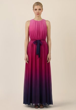 Frau steht mit Blick nach vorne, trägt ein ärmelloses, bodenlanges Ombre-Kleid in Pink- und Violetttönen mit einem gebundenen Gürtel in der Taille.
