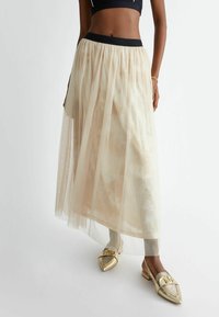 Gonna lunga in tulle beige con una trama trasparente e una vita nera a contrasto. Abbinata a scarpe dorate con punta affilata e fibbia decorativa.