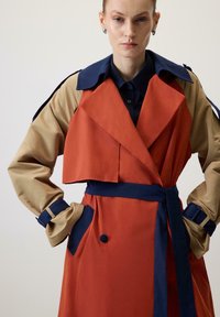 Abrigo tipo trench que presenta una mezcla de tejido naranja y beige, diseño estructurado, amplio cuello, cintura con cinturón y acentos en azul marino en contraste.