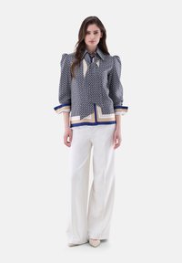 Bluse mit Marine-Muster, Puffärmeln, kontrastierendem Kragen und goldenen Akzenten, kombiniert mit weißen weiten Hosen und beigen Schuhen.