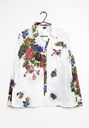 Biała koszula z długim rękawem z kolorowym kwiatowym wzorem w odcieniach czerwieni, niebieskiego, zieleni i brązu, z kieszenią na piersi, zawieszona na czarnym wieszaku.