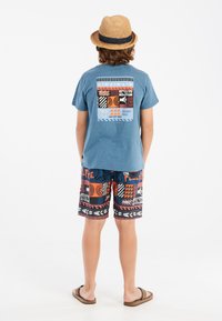 Camiseta de algodón azul con un gráfico en la parte trasera con un patrón de olas y motivos de surf; combinada con pantalones cortos de baño de patrones llamativos en naranja y negro.