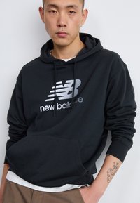 Črna bombažna kapuca z žepom na sprednji strani in kapuco na vrvici. Značilnost srebrnega logotipa New Balance na prsih. Enostavna tekstura.