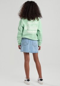 Hellgrüner Sweatshirt mit weißem Grafiktext auf dem Rücken, kombiniert mit einem Jeansrock und weißen Sneakers mit schwarzen Akzenten.