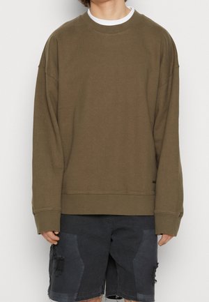 Osoba ubrana w oliwkowozieloną, długorękawową bluzę nałożoną na białą koszulę, w połączeniu z czarnymi, podartymi szortami dżinsowymi.