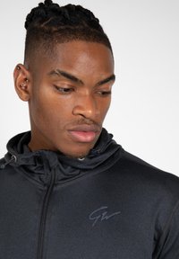 Zwarte zip-up hoodie met een hoge kraag, trekkoordaccenten en een subtiele logo op de borst. Gladde stof met een getailleerd ontwerp.