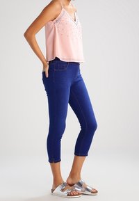 Ljusrosa ärmlös topp med V-ringning och dekorerad med silvernitar, i kombination med marinblå caprijeans och metalliska sandaler.