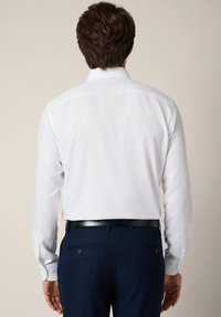 Biała koszula z długim rękawem zapinana na guziki przy mankietach, o gładkiej fakturze, zestawiona z ciemnoniebieskimi spodeniami na miarę z bocznymi kieszeniami i czarnym paskiem.