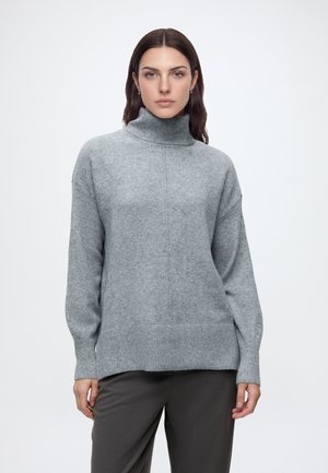Femme portant un pull à col roulé gris et un pantalon sombre, debout devant un fond clair uni, regardant devant avec une expression neutre.