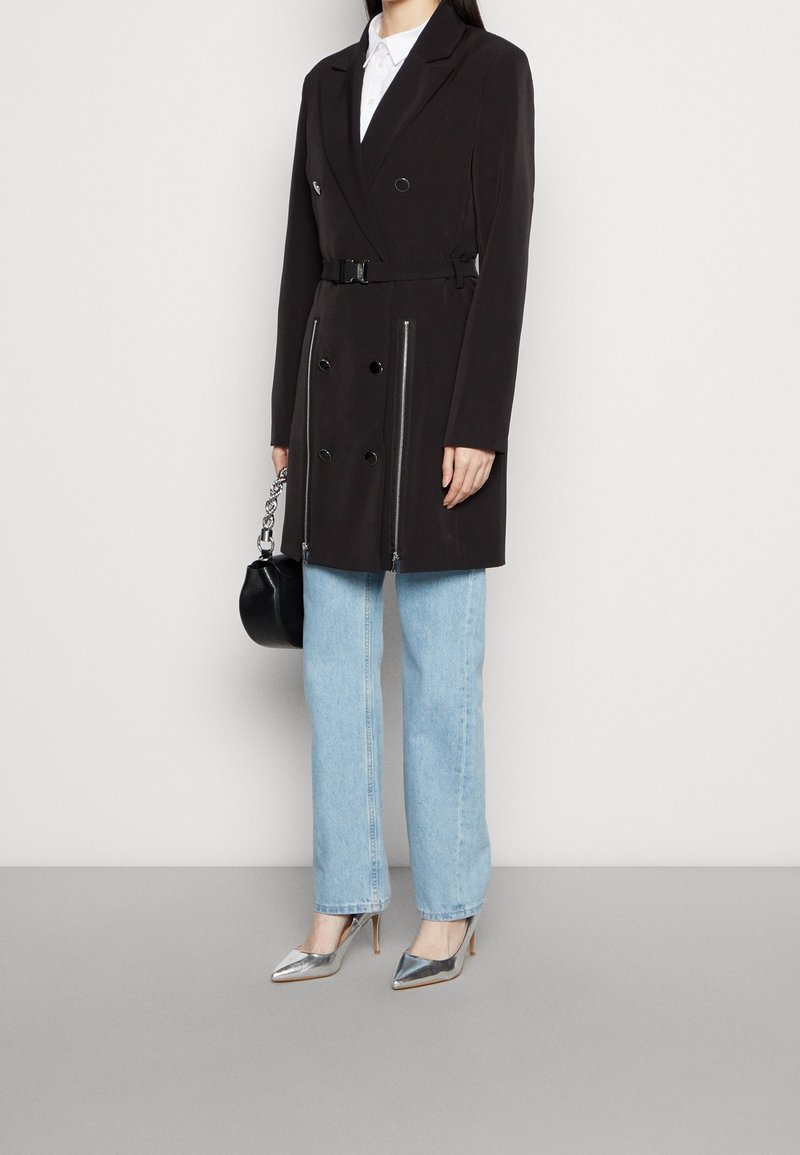 Manteau noir croisé à double boutonnage avec une ceinture, détails de fermeture éclair argentés, porté sur une chemise blanche, associé à un jean bleu clair et des talons argentés.