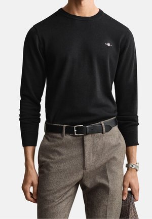 Homme portant un pull noir à manches longues avec un petit logo, un pantalon marron en chevrons, une ceinture noire et une montre-bracelet argentée, tenant un tissu dans la main.