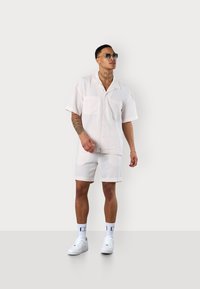 Weißes kurzärmliges Hemd mit Kragen und passender Shorts mit einem dezenten karierter Muster, kombiniert mit weißen Turnschuhen und Sonnenbrille.