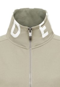 Svetlobéžová bunda na zips s vysokým golierom. Na golieri sú biele blokové písmená a vnútorná podšívka má textúru. Hladký povrch látky.