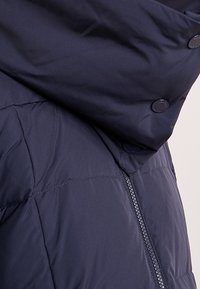 Mörkblå vadderad jacka med hög krage som har tryckknappar och en synlig vertikal dragkedja på sidan.