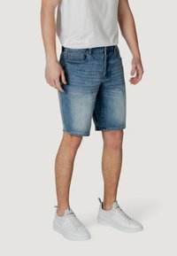 Pantalones cortos de mezclilla azul con acabado deslavado, que llegan a la mitad del muslo. Presentan un cierre con botón y cremallera, cinco bolsillos y presillas para cinturón. Zapatillas de tonos neutros.