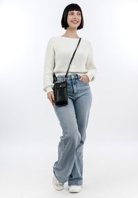 Black leather crossbody phone bag with a silver button, worn over a white ribbed sweater and light blue high-waisted jeans. Sneakers visible.