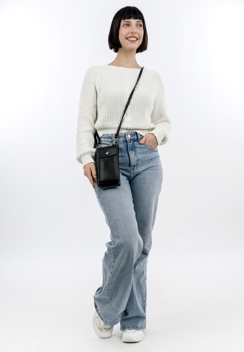 Black leather crossbody phone bag with a silver button, worn over a white ribbed sweater and light blue high-waisted jeans. Sneakers visible.