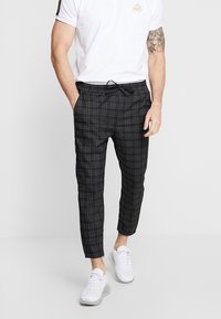 Man i vit t-shirt och svarta rutiga byxor står med händerna i fickorna, iförd vita sneakers mot en enfärgad bakgrund.