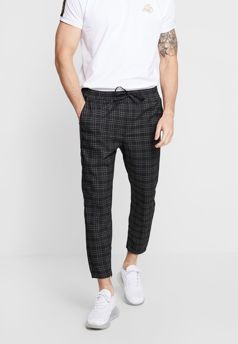 Man i vit t-shirt och svarta rutiga byxor står med händerna i fickorna, iförd vita sneakers mot en enfärgad bakgrund.