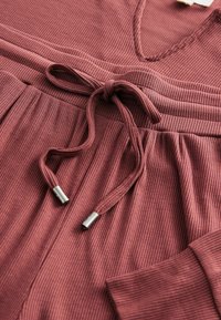 Mörkröd ribbad loungewear-set med V-ringning, dragsko i midjan och aglets i silverfärgad metall. Har avslappnade ärmar och fickor.