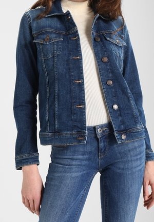 Kvinde iført en blå denimjakke over en hvid ribbet rullekravetrøje og blå denimjeans mod en ensfarvet baggrund.