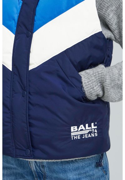 Marineblaues Steppweste mit weißem und hellblauem Zickzackmuster, mit einem in Weiß gestickten Logo und einem grauen Strickpullover, der darunter sichtbar ist.