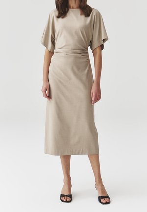 Beżowa sukienka midi z miękkiego materiału, z okrągłym dekoltem, luźnymi rękawami i dopasowaną talią, uzupełniona eleganckimi czarnymi sandałami.