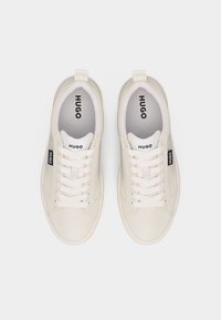 Poros beveik baltų Hugo sportbačių su baltais raišteliais, juodais Hugo logotipo ženkliukais šonuose, o liežuvėlyje ir vidpadžiuose pažymėta Hugo, žiūrint iš viršaus.