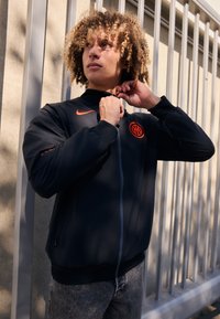 Crna jakna na zip s visokim ovratnikom, s narančastim Nike logotipom i okruglim grbom na prsima. Model prilagođava ovratnik, a tkanina je teksturirana.