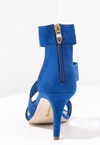 Sandal con tacco alto in morbido camoscio blu, con cerniera posteriore e cinturino alla caviglia, vista da dietro su una superficie bianca.