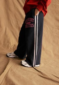 Pantaloni sportivi blu marina con strisce laterali bianche e testo rosa "Superdry Athletics", abbinati a una felpa rossa e scarpe sportive grigie.