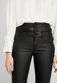 Hoge taille zwarte jeans met een gladde, glanzende textuur, voorzien van een vijfknops sluiting en zijzakken. Aangevuld met gestikte details.