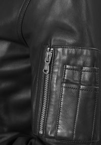 Bereich mit schwarzer Lederjacke, der eine mit Reißverschluss versehenen Tasche mit gerippter Textur und glatter Oberfläche zeigt. Auffälliges silbernes Reißverschluss-Hardwaredetail.