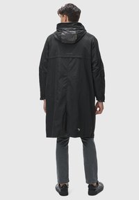 Svart lätt regnrock med huva, långa ärmar och avslappnad passform. Har en ventil på baksidan och en liten varumärkesemblem på fållen.