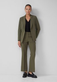 Olivgrüner, taillierter Blazer mit einem einzigen Knopf und Revers, kombiniert mit passenden schmal geschnittenen Hosen und schwarzen Lacklederschuhen.