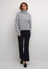 Lichtgrijze, gebreide coltrui met gewatteerde diamanten patronen, gecombineerd met donkere, uitlopende jeans en zwarte puntige ballerina's.