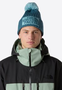 Berretto lavorato a maglia in teal scuro con pom-pom teal chiaro e patch con logo. Il modello indossa una giacca impermeabile nera e verde menta con colletto alto.
