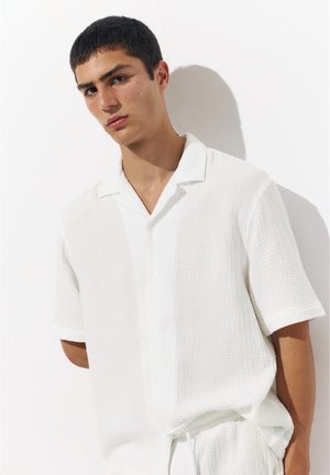 Jeune homme aux cheveux courts et foncés portant une chemise blanche texturée à manches courtes ample et un pantalon assorti devant un mur blanc.
