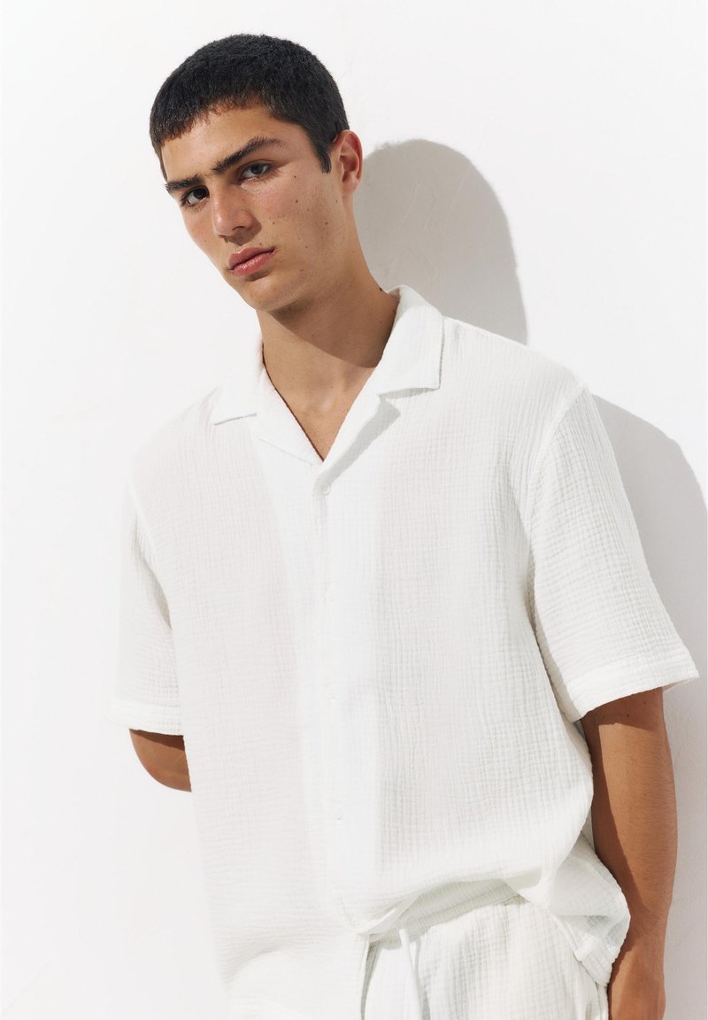 Jeune homme aux cheveux courts et foncés portant une chemise blanche texturée à manches courtes ample et un pantalon assorti devant un mur blanc.