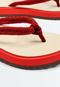 Gros plan d'une sandale beige et rouge avec des brides en tissu rouge texturé et une semelle épaisse rouge et marron.
