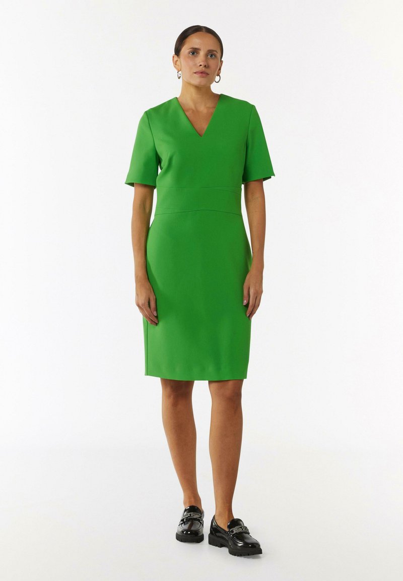 Zielona sukienka z dekoltem w serek i krótkimi rękawami, dopasowana w talii, o długości do kolan, uszyta z gładkiego materiału. W połączeniu z czarnymi mokasynami.