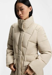 Beige puffjacka med hög krage, med diamantformad vaddering, knappfäste framtill och sidofickor. Slät textur.