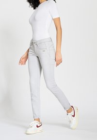 Jeans skinny gris claro con una textura suave, bolsillos delanteros y cierre de cremallera. Llevados con zapatillas blancas con acentos rojos.