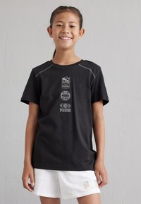 Camiseta negra de manga corta que presenta tres logotipos grises de Puma en el frente, con costuras reflectantes en los hombros. Combinada con pantalones cortos blancos.