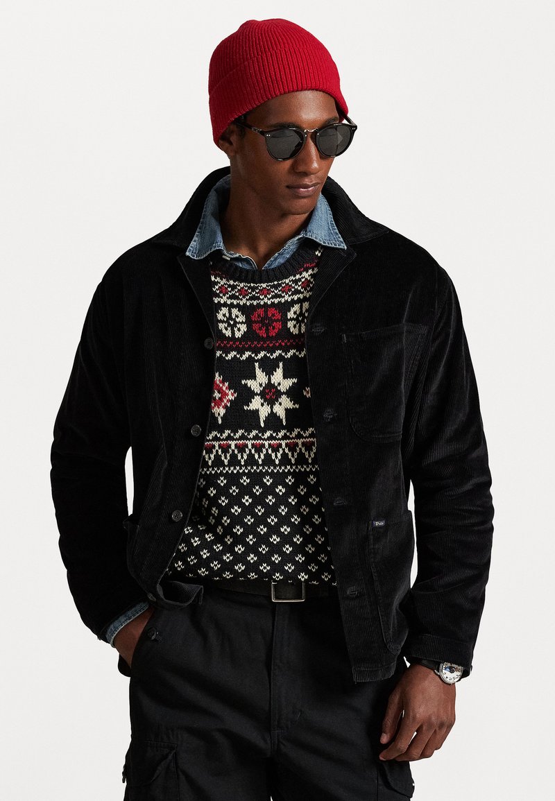 Black corduroy jacket over a knitted sweater with red and white geometric patterns, paired with a denim shirt, black cargo pants, and a red beanie.