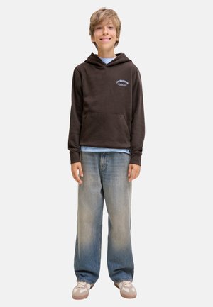 Un garçon souriant et debout, portant un sweat à capuche marron foncé, un jean bleu délavé et des baskets beiges sur un fond uni.