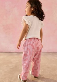 Haut à côtes blanches avec des manches volantées, associé à un pantalon cargo rose avec des motifs abstraits. Mis en valeur sur un fond rose doux.