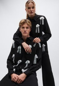 Μαύρα πουκάμισα με κουμπιά, με σχέδια λευκών δαντελωτών φιόγκων. Το ύφασμα έχει απαλή υφή. Ο σχεδιασμός περιλαμβάνει πολλούς φιόγκους μπροστά.