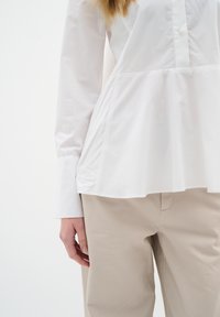 White long-sleeve blouse with a pleated design at the waist, paired with beige trousers. The fabric appears smooth and lightweight.