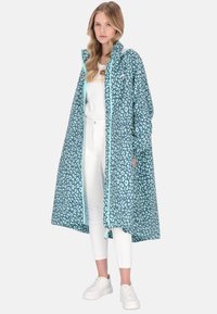 Haină impermeabilă albastră cu imprimeu leopard, design lung, fermoar de culoare teal deschis și manșete elastice. Asortată cu pantaloni albi și adidași.